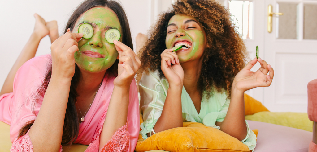 two woman smiling with green facial mask on skin playing with cucumber slices 