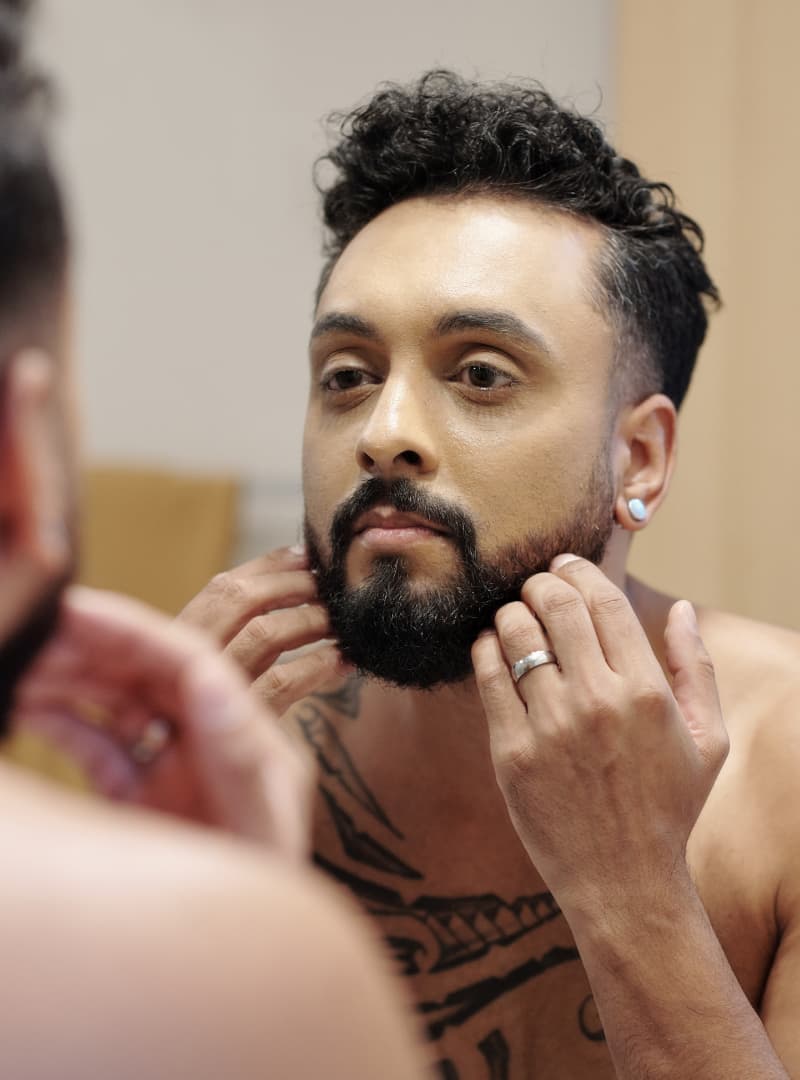Man with a beard and tattoos examining his beard in a mirror.
